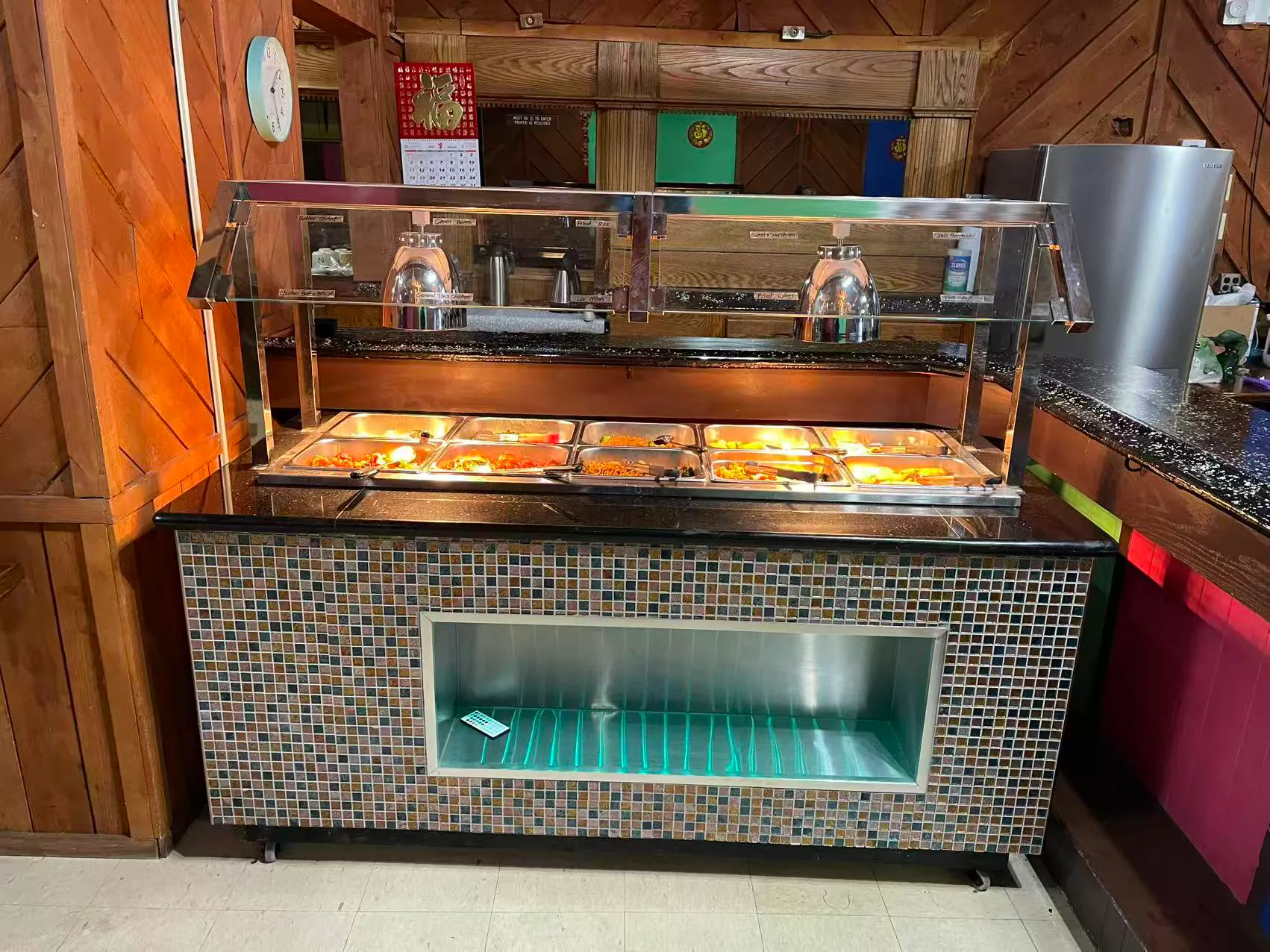 A buffet station with a glass sneeze guard, displaying several trays of hot food against a backdrop of wooden walls.